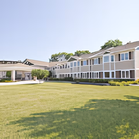 Exterior view of a two-story senior living facility building with multiple windows, surrounded by a well-maintained green lawn and some trees in the background under a clear sky.
