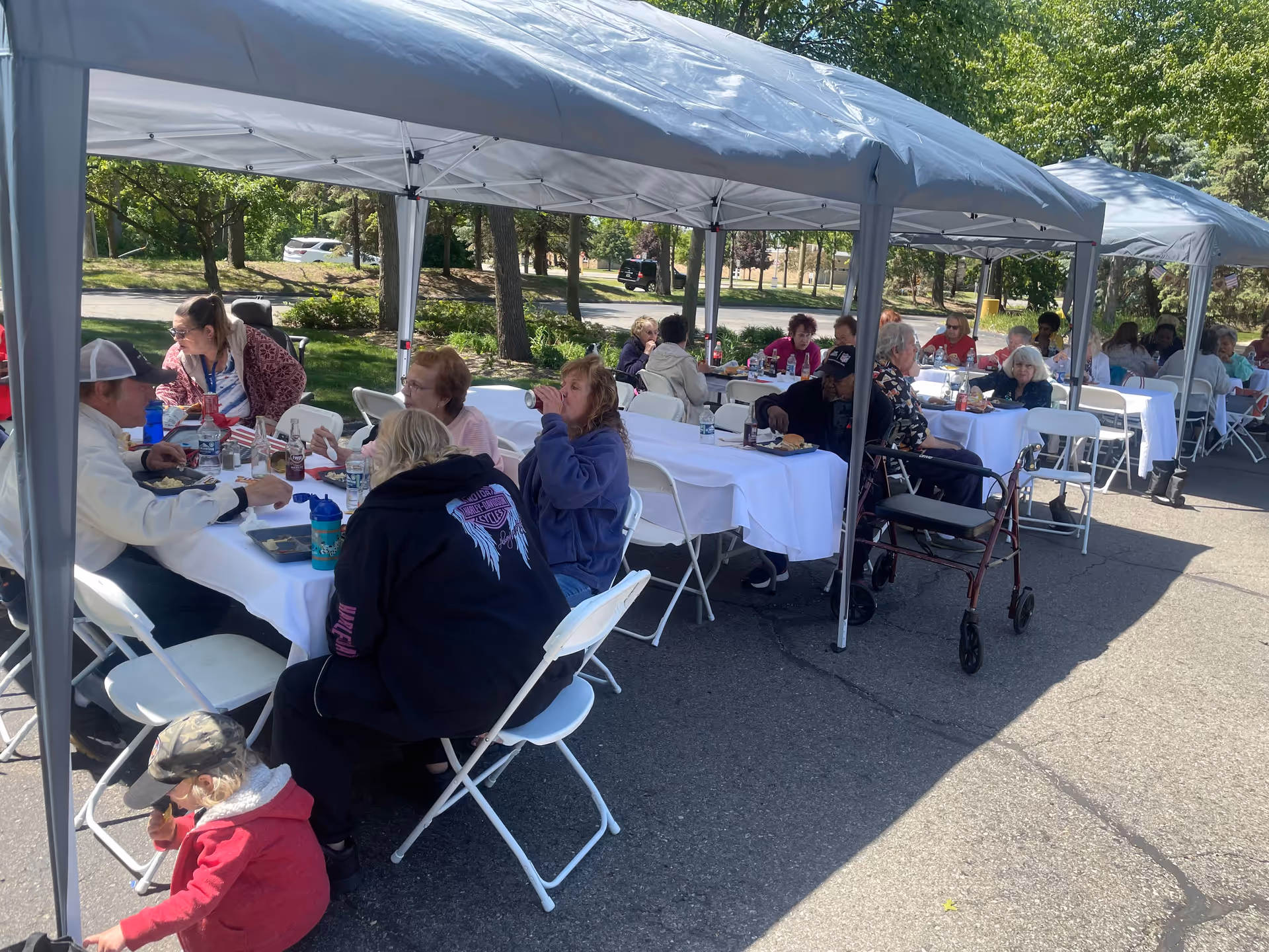 A group of seniors seated at long white-clothed tables under outdoor canopy tents, eating and socializing.