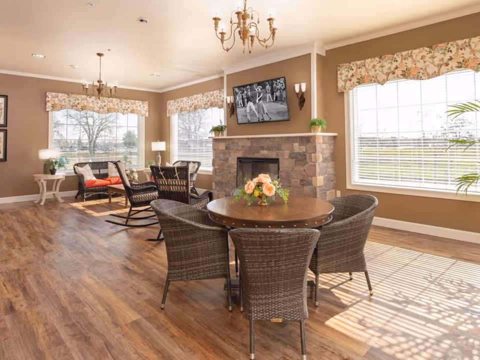 A bright and cozy living room area with large windows covered by floral valances allowing natural light to fill the space. The room features a stone fireplace with a black and white photo mounted above it, a round wooden table with four wicker chairs, and additional seating including a rocking chair and a cushioned bench. The floor is wooden, and there are plants and lamps placed around the room.