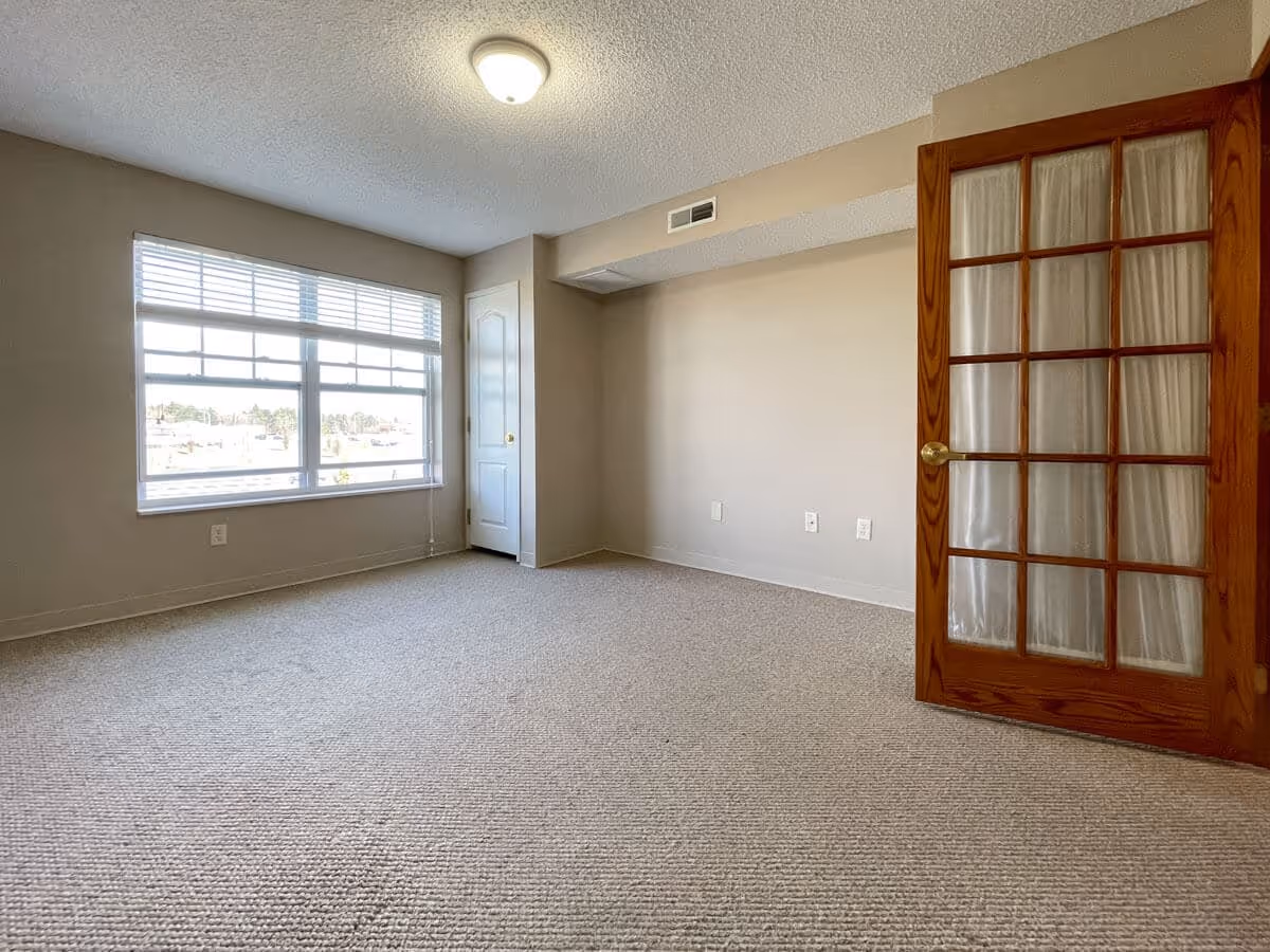Empty carpeted bedroom with a large window, small closet, and a wooden French door with glass panes.