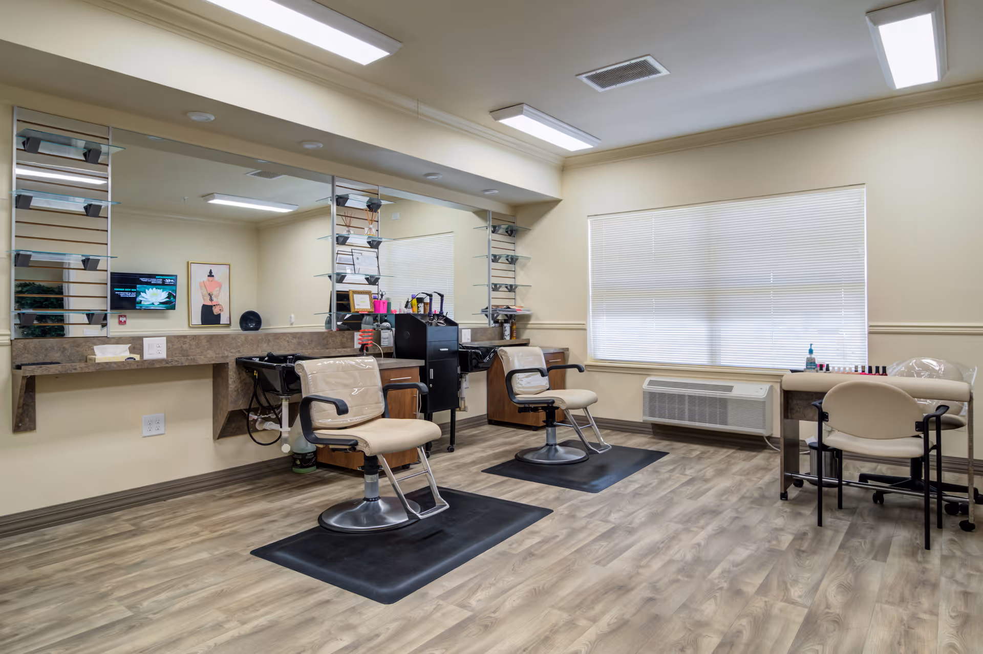 Interior view of a salon area in a senior living facility with two salon chairs in front of a large mirror, a manicure table with a chair, and various salon equipment. The room has wood flooring, beige walls, and a large window with blinds.