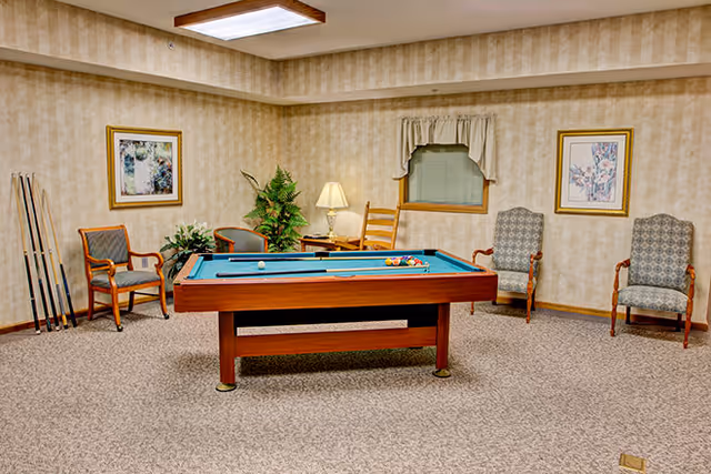 A recreational room with a pool table in the center, surrounded by four chairs, two framed floral paintings on the walls, a small window with a valance, a floor lamp, and a plant in the corner. Pool cues are standing against the wall on the left side.