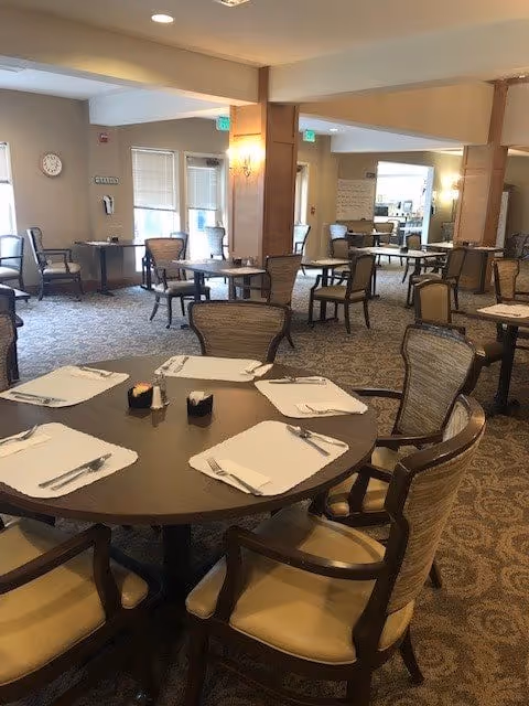 A spacious dining room with multiple round and rectangular tables set with placemats, napkins, and silverware. The room has carpeted floors, beige walls, and several chairs around each table. There are windows with blinds, wall sconces, and a clock on the wall.