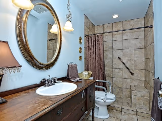 A bathroom with a wooden vanity featuring a white sink and a large oval mirror above it. There is a lamp on the left side of the vanity and a towel hanging on the wall. The shower area has beige tiles, a built-in bench, a grab bar, and a brown shower curtain. A toilet with armrests is positioned next to the shower.
