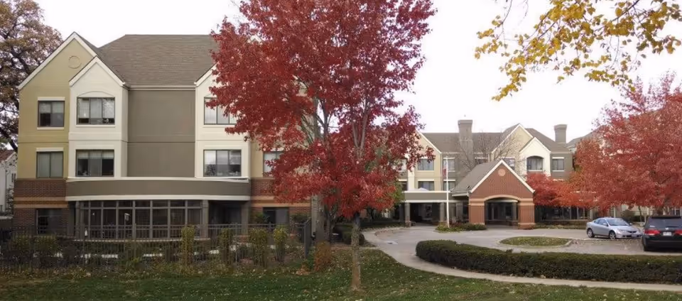 Front exterior of a three-story retirement campus building with red autumn trees, a circular driveway, and parked cars.