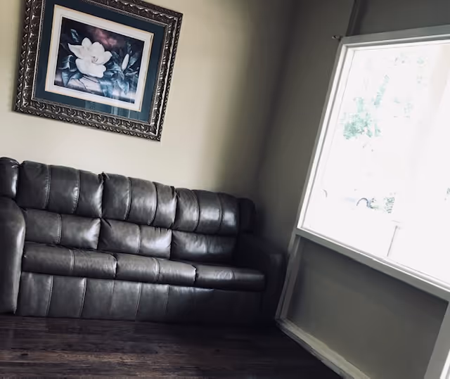 A corner of a room featuring a dark gray leather couch against a beige wall. Above the couch is a framed painting of white flowers with a dark background. To the right, there is a large window letting in natural light, showing some greenery outside. The floor is wooden with a dark finish.