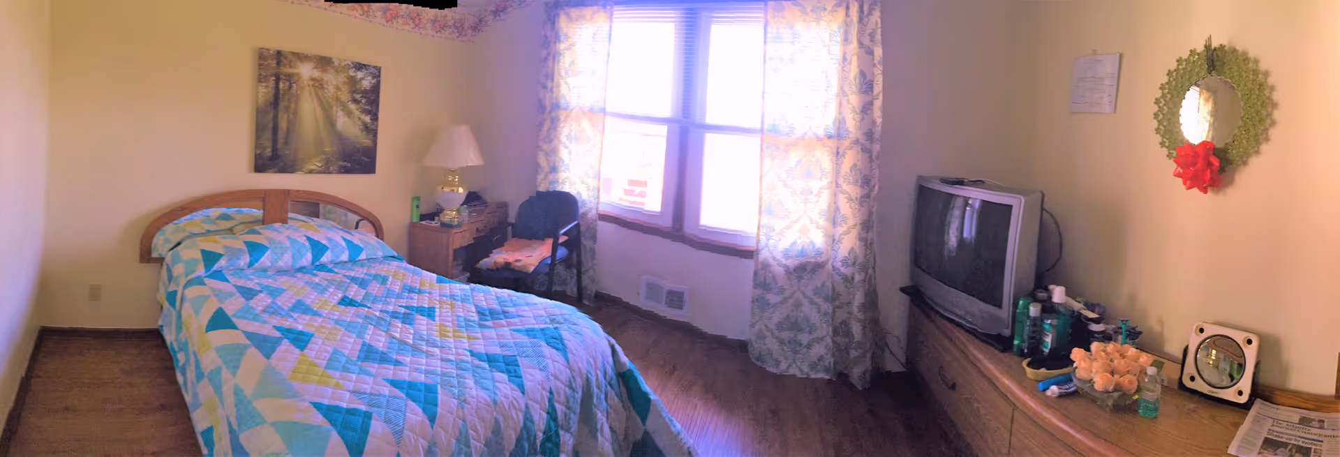 A small bedroom with a single bed covered in a colorful quilt. There is a wooden nightstand with a lamp and some items next to the bed. A chair with a folded blanket is placed near a window with patterned curtains. Across from the bed is a wooden dresser with a TV, various personal care items, a small mirror, and a vase of flowers. A decorative wreath with a red bow hangs on the wall above the dresser.