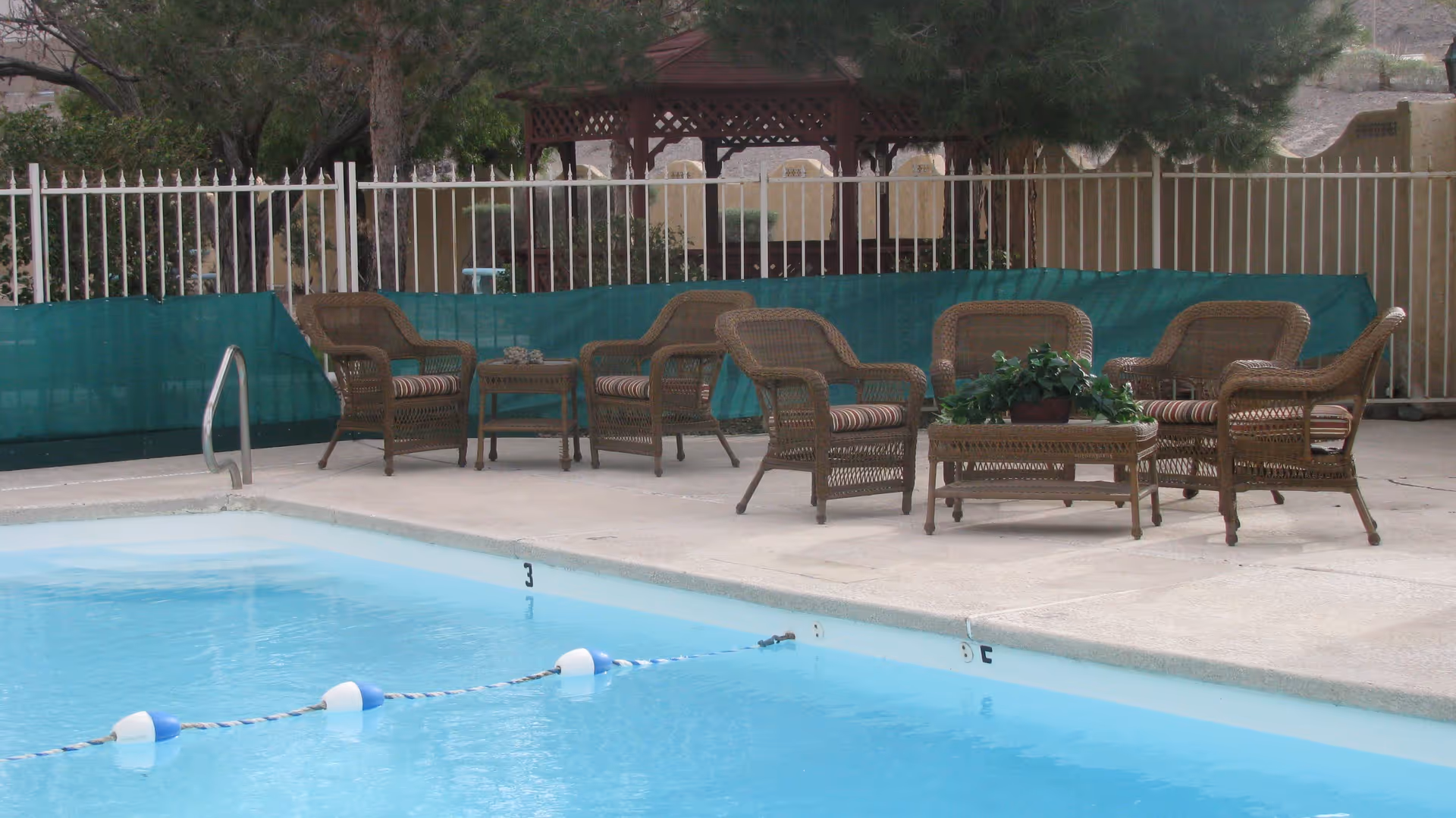 Outdoor swimming pool with a concrete deck lined with wicker chairs and tables beside a metal fence.