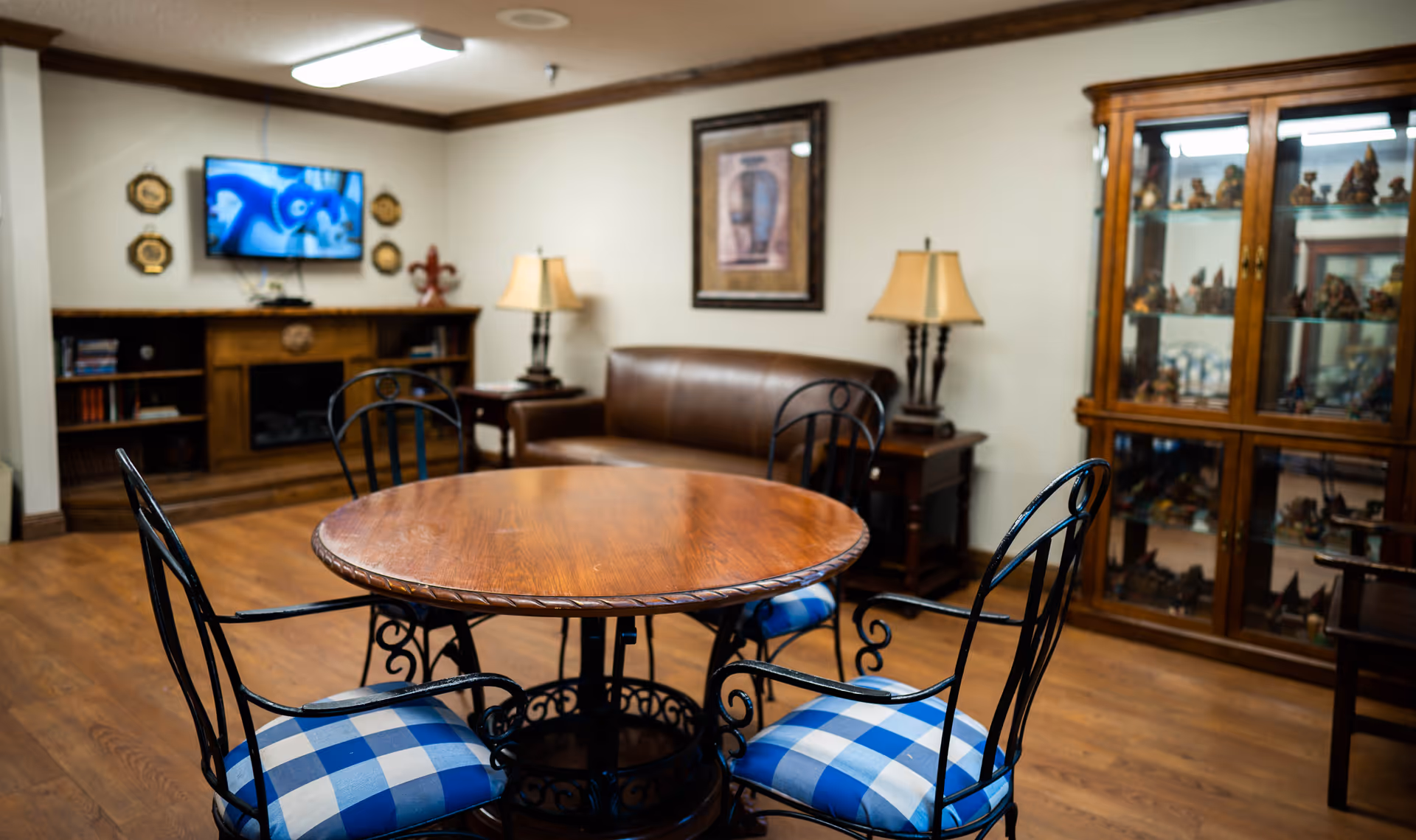 A cozy living room with a round wooden table surrounded by four black metal chairs with blue and white checkered cushions. In the background, there is a brown leather sofa, two side tables with lamps, a wooden cabinet with glass doors displaying various figurines, and a wall-mounted TV showing an animated movie. The room has wooden flooring and neutral-colored walls with framed artwork.