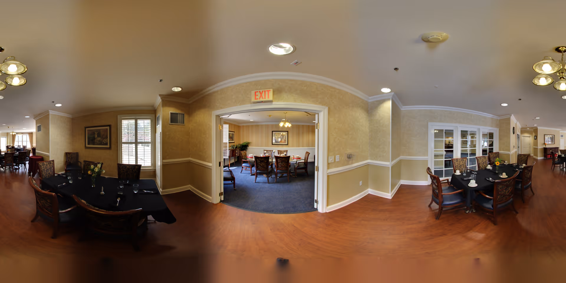 Interior view of a senior living facility dining area with wooden floors and beige walls. There are multiple dining tables covered with black tablecloths, set with glasses and napkins. The room has traditional-style chairs and is well-lit with ceiling lights and natural light from windows. A doorway leads to another room with additional dining tables and chairs. The overall atmosphere is warm and inviting.