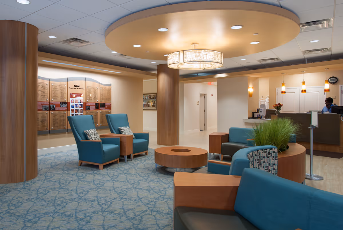 A modern and comfortable senior living facility lobby with teal upholstered chairs and sofas arranged around a circular wooden coffee table. The ceiling features a large circular light fixture, and there are wooden columns and a blue patterned carpet. In the background, a reception desk with a staff member is visible, along with wall-mounted plaques and pendant lights.