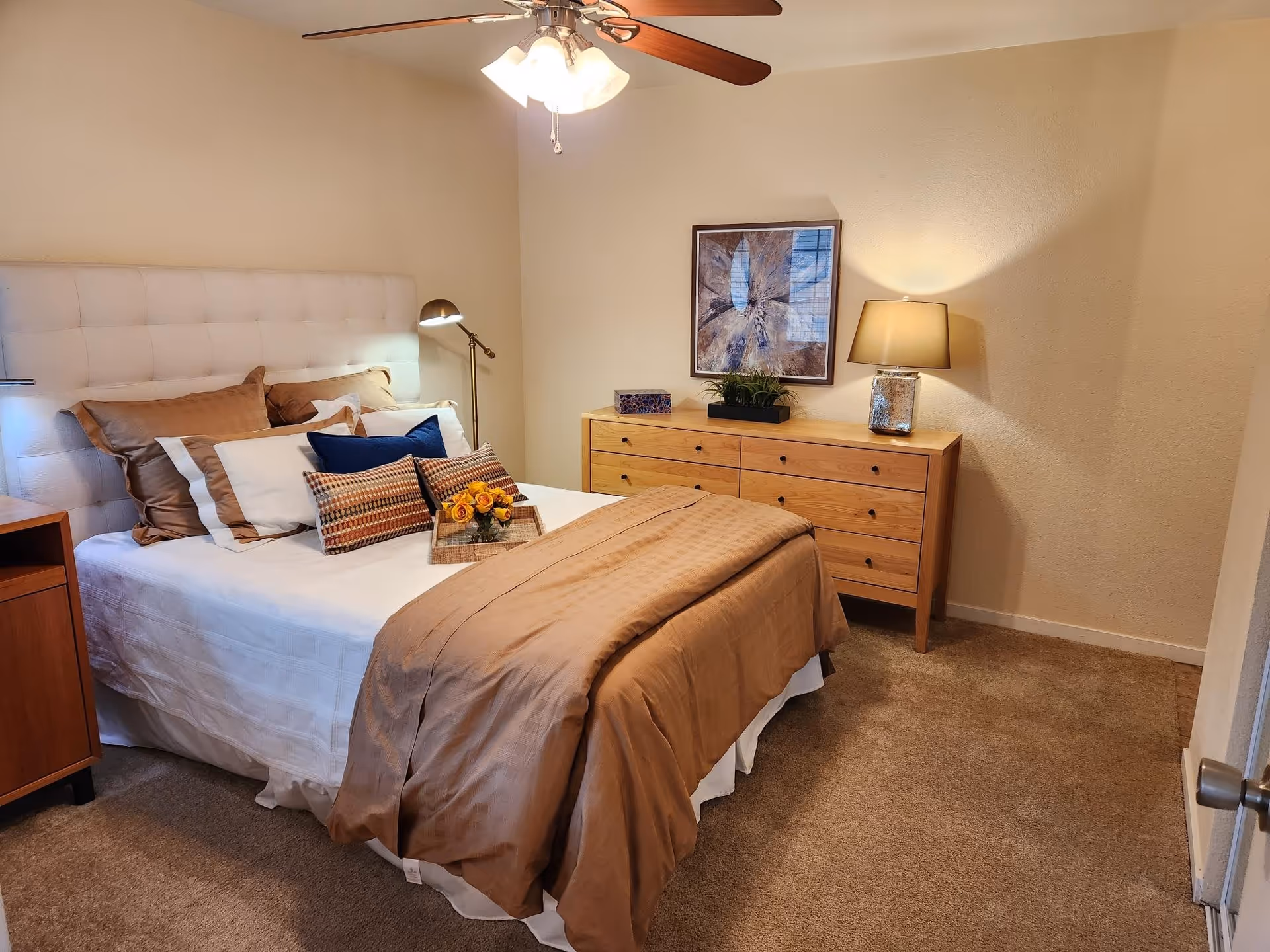 A cozy bedroom with a neatly made bed featuring multiple pillows and a brown comforter. There is a wooden dresser with a lamp, a plant, and a decorative box on top. A framed artwork hangs on the wall above the dresser. The room has beige walls and carpeted flooring, with a ceiling fan and light fixture above.