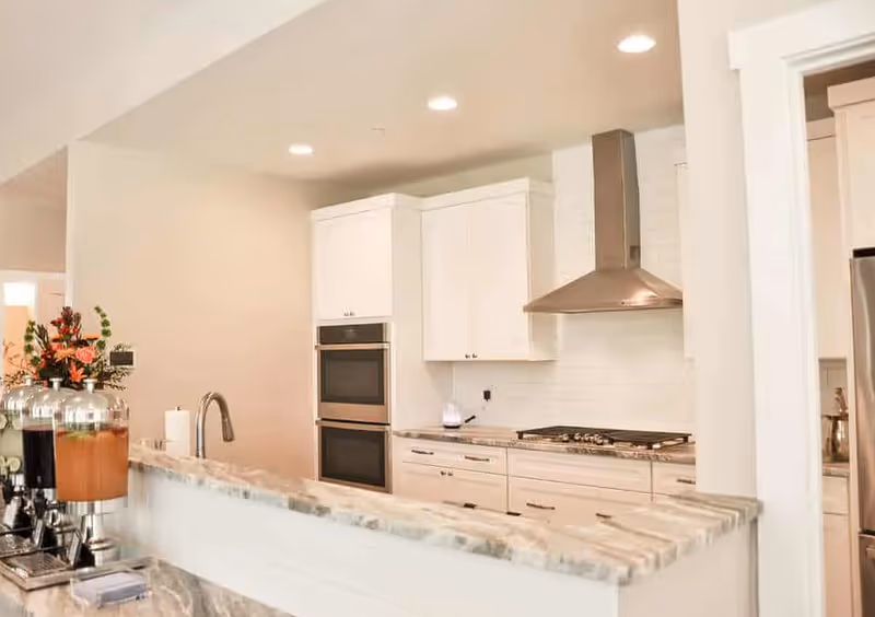 Modern kitchen with white cabinets, a marble countertop, a built-in double oven, a stainless steel range hood, and a gas stove. There are beverage dispensers and a flower arrangement on the counter.