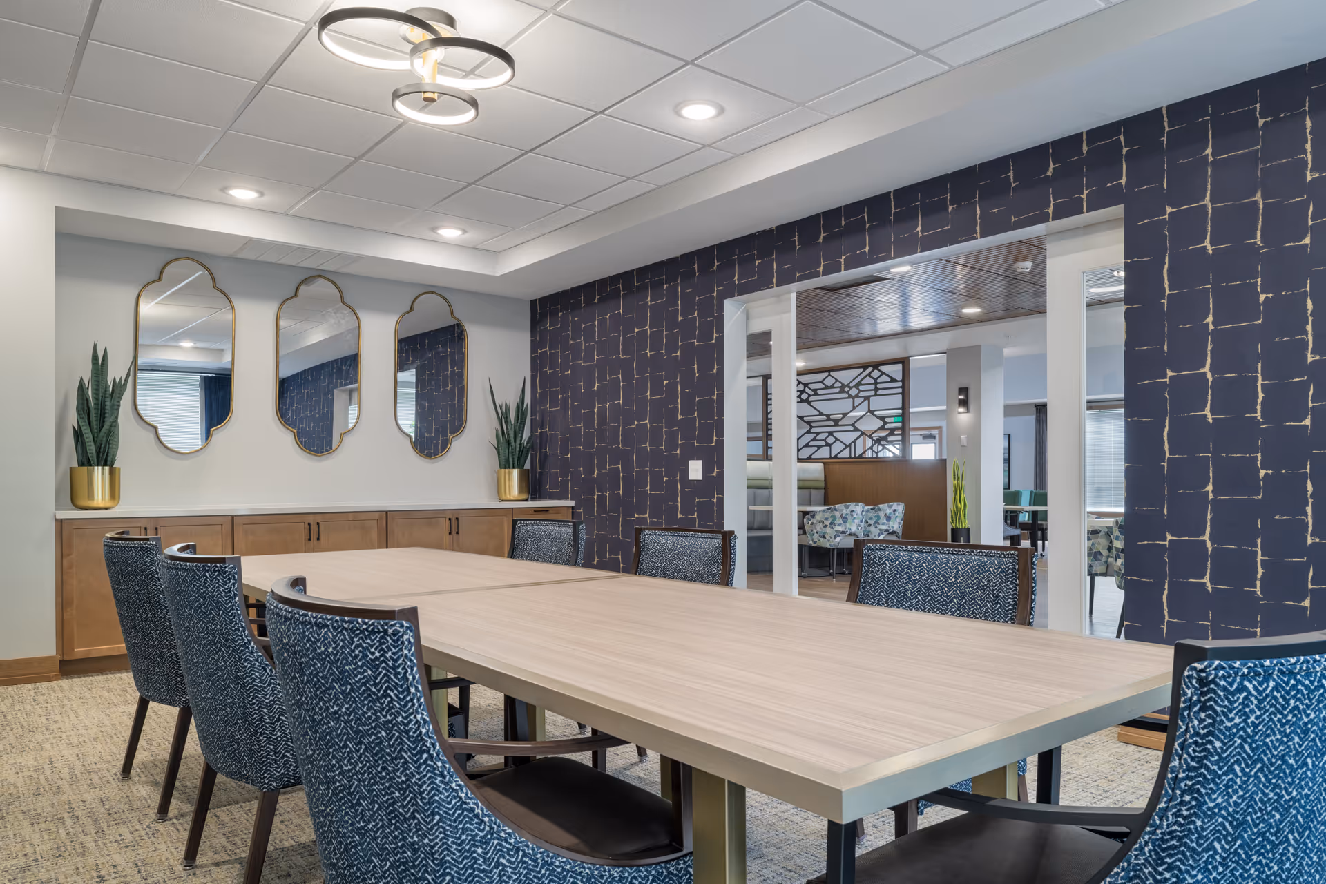 A modern dining room with a long light wood table surrounded by eight blue patterned chairs. The room features a dark blue accent wall with a gold grid pattern, three decorative mirrors with gold frames on a white wall, and two potted plants in gold pots on a wooden sideboard. The ceiling has recessed lighting and a contemporary circular light fixture.