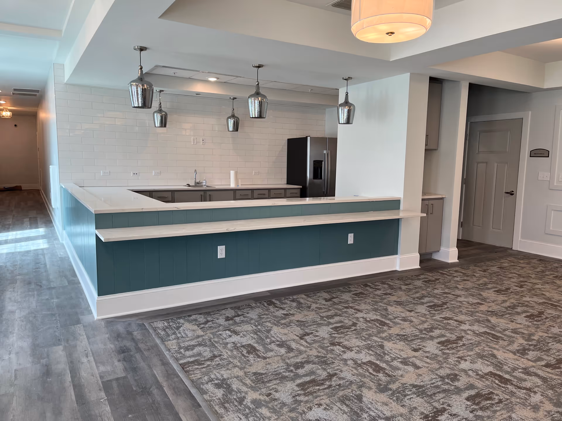 Interior view of a modern kitchen area with a U-shaped counter featuring a teal base and white countertop. The kitchen has gray cabinets, a stainless steel refrigerator, a sink, and four metallic pendant lights hanging from the ceiling. The floor is a combination of wood and patterned carpet, and there is a hallway visible to the left.