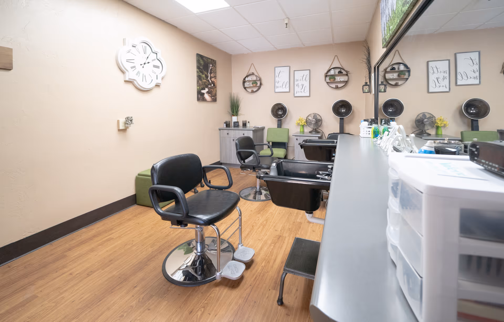 Interior view of a hair salon area in a care center with black salon chairs, hair drying stations, a large mirror, wooden flooring, and wall decorations including a white clock and framed quotes.