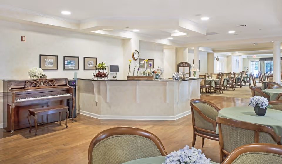 A spacious dining room in a senior living facility with multiple tables covered in green tablecloths and decorated with small flower arrangements. There is a wooden piano with a bench on the left side, and a counter area with decorative items and a clock on the wall behind it. The room has wooden flooring and soft lighting from ceiling fixtures.