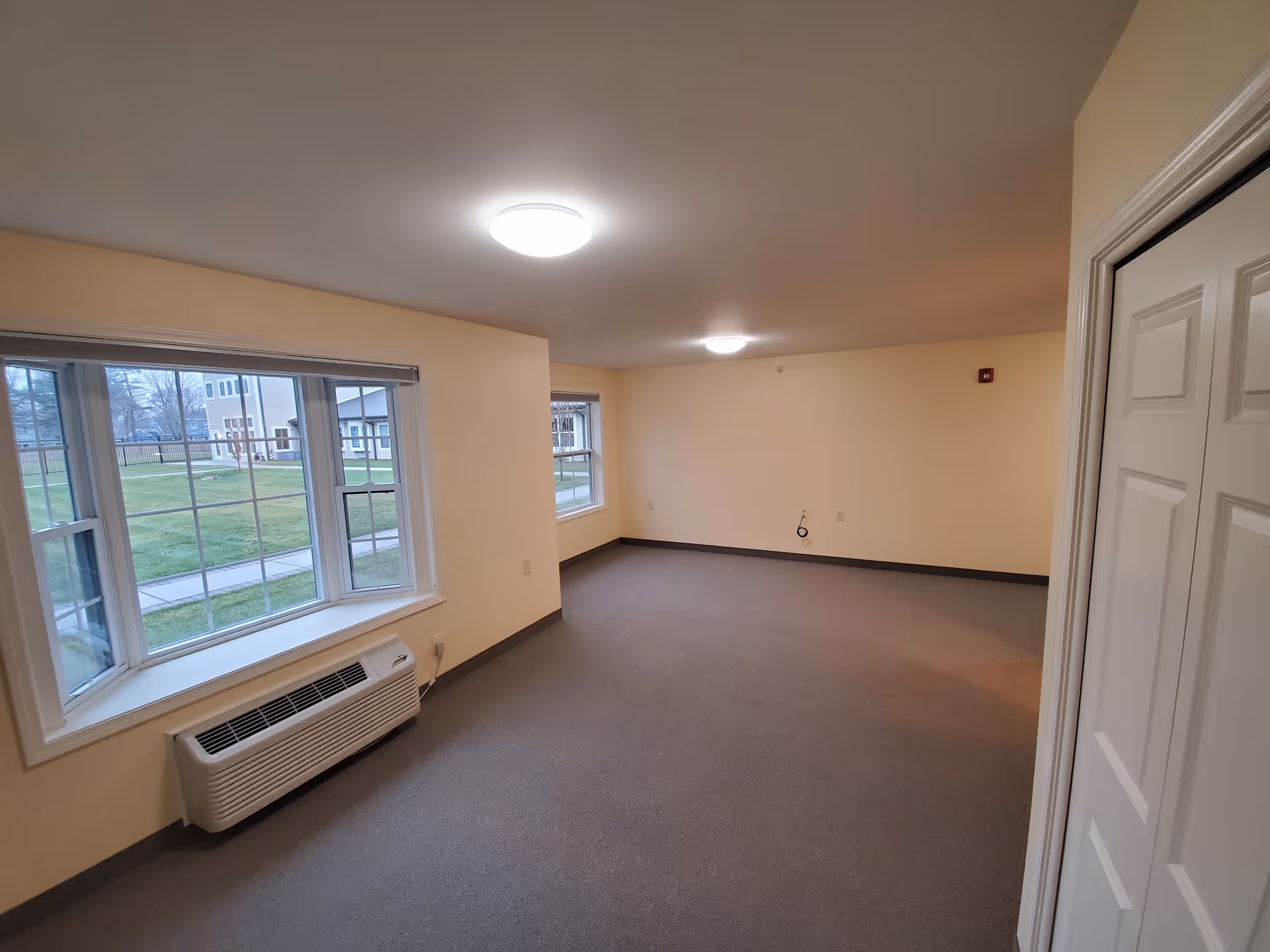 Empty room with beige walls and gray carpet, featuring two large windows with white frames letting in natural light. There is a white heating/cooling unit below one window and a closed white closet door on the right side. The ceiling has two round flush-mounted lights.