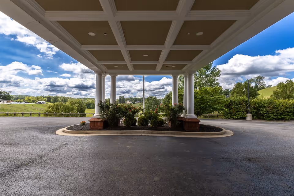 Covered driveway entrance with white columns and a landscaped island with bushes and flowers in the center, set against a backdrop of green trees and a partly cloudy blue sky.