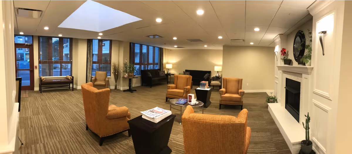 A spacious and well-lit common area in Brooklyn Boulevard ALP featuring multiple comfortable armchairs arranged around small tables, a fireplace with decorative plants and a clock above it, large windows with blinds, and a bench near the entrance door. The room has a carpeted floor and recessed ceiling lights.