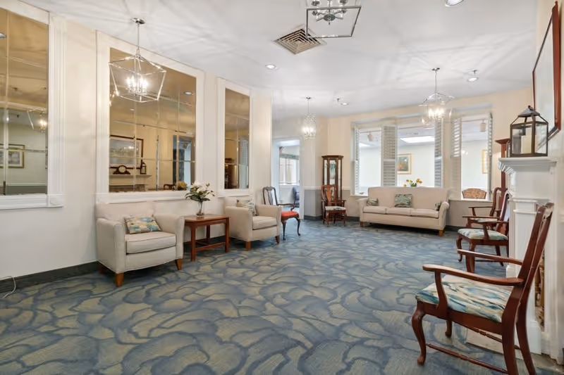 A spacious senior living common area with blue patterned carpet, beige armchairs, a beige sofa, wooden chairs with patterned cushions, a wooden side table with a flower vase, large mirrors on one wall, and pendant lights hanging from the ceiling.