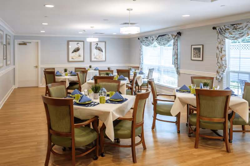 A bright dining room with several tables covered in white tablecloths, each set with blue napkins, green mugs, and glassware. The room features wooden chairs with green cushions, large windows with floral curtains, and framed bird artwork on the walls. The floor is wooden, and ceiling lights provide illumination.