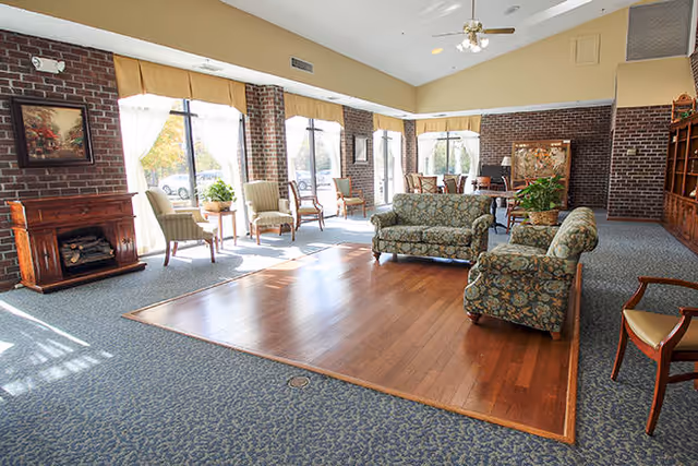 Sunlit communal lounge with upholstered sofas and chairs around a wooden seating area and large windows.