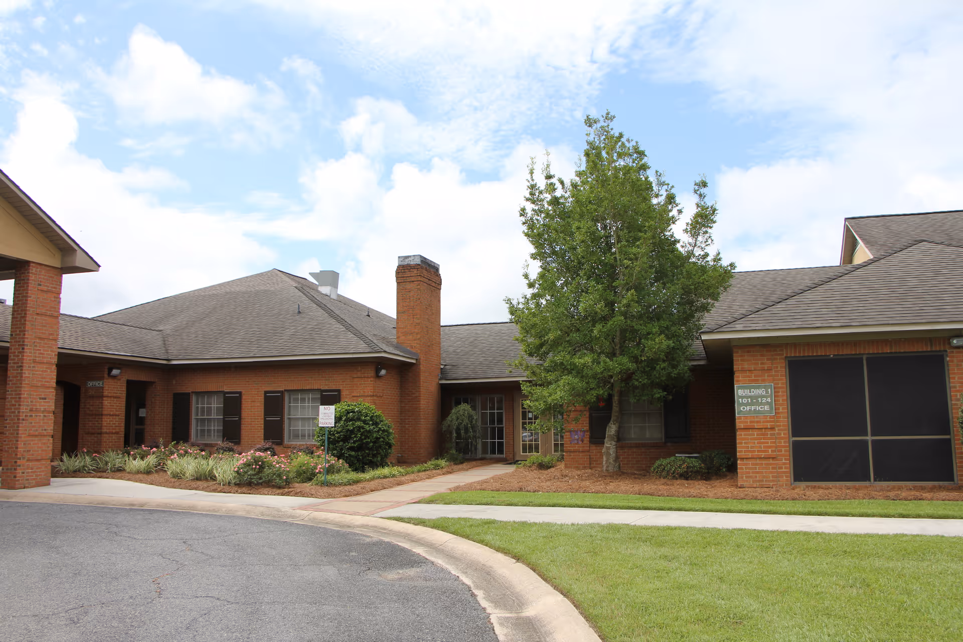 Exterior view of a single-story brick building with a gray shingled roof, a chimney, and several windows with dark shutters. There is a tree and landscaped bushes in front of the building, along with a curved driveway and a sidewalk. A green sign on the building indicates 'Building 1 101-124 Office'. The sky is partly cloudy.
