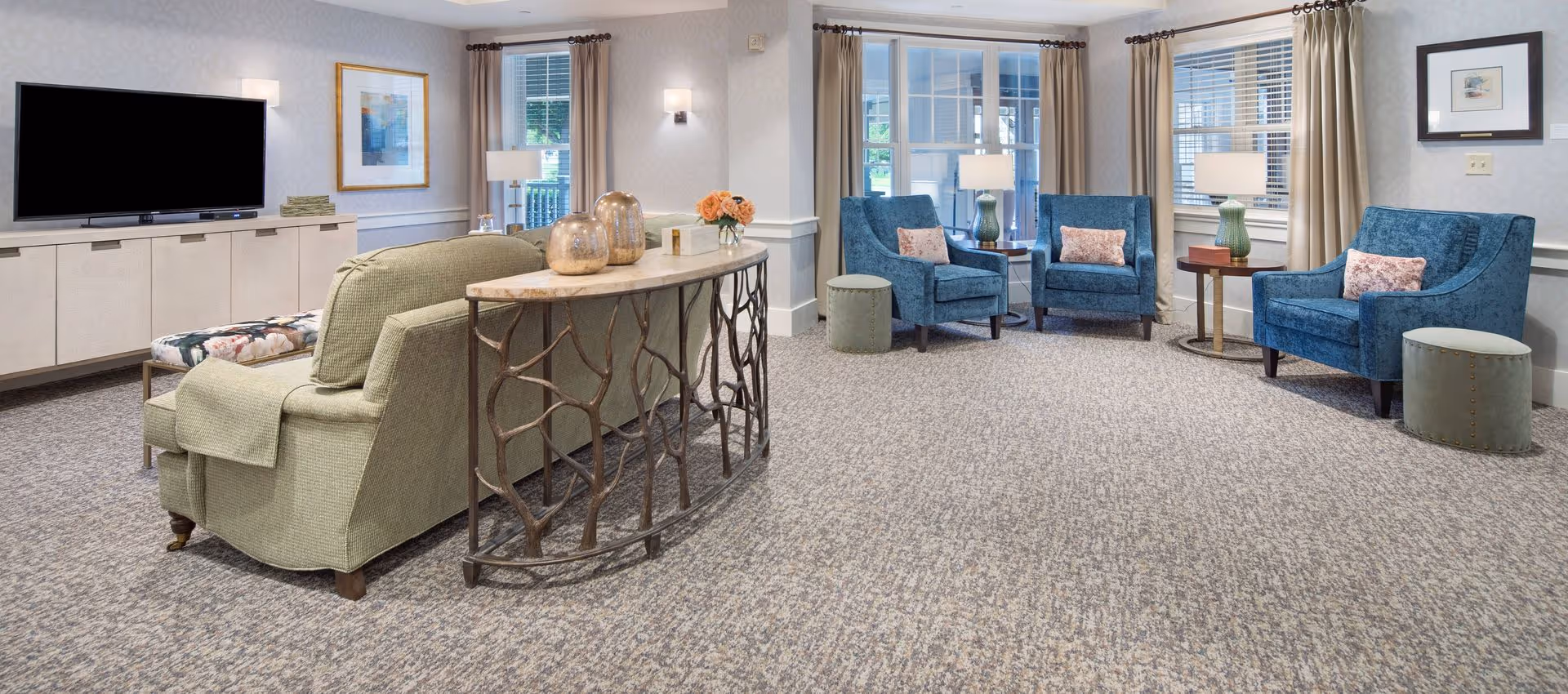 Bright, carpeted communal living room with sofas and armchairs arranged around a TV and a decorative console table.