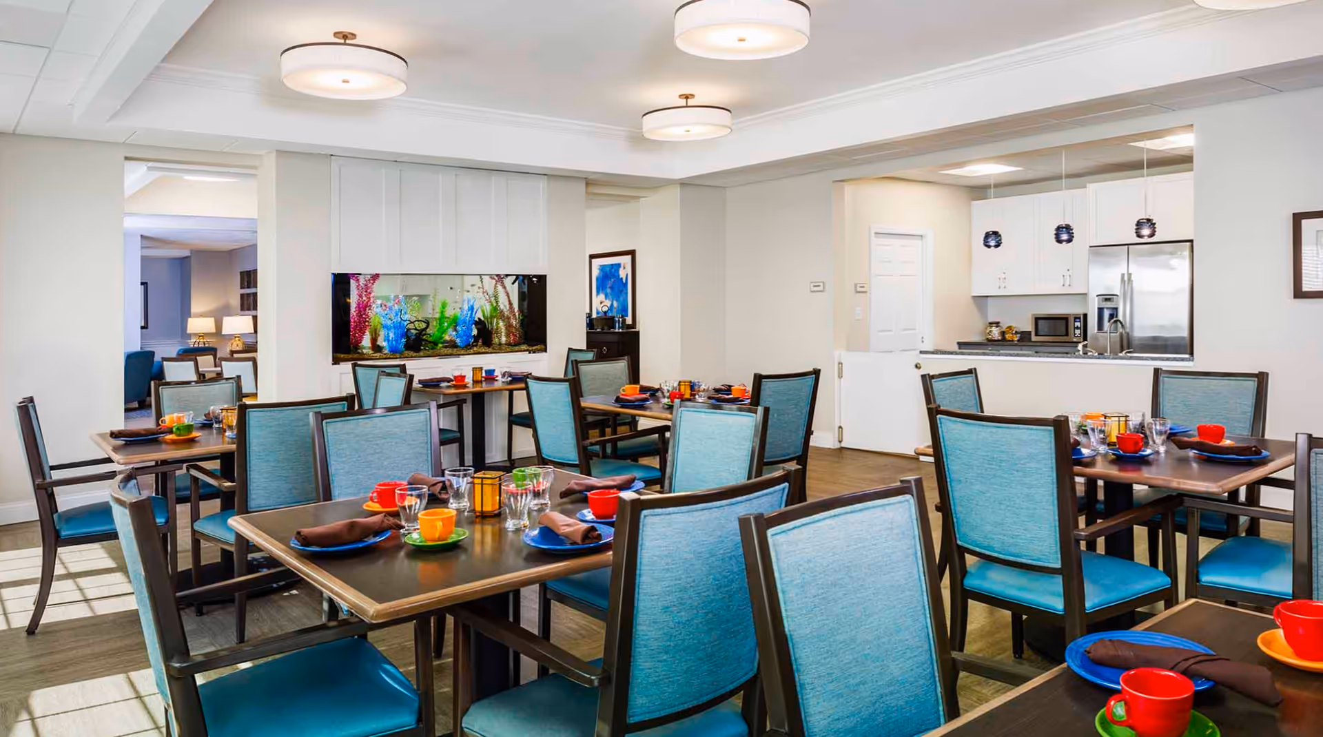 A bright and clean dining room with multiple tables set with colorful cups, plates, and napkins. The room features teal upholstered chairs, a large built-in aquarium, and a kitchen area with white cabinets and stainless steel appliances in the background.