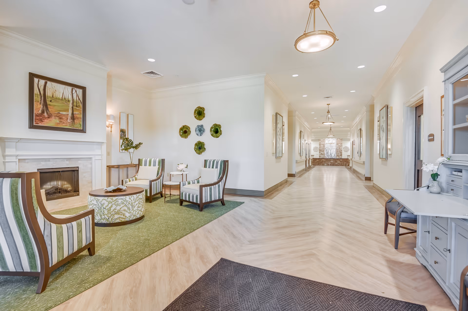 Bright senior living facility common area with striped armchairs around a coffee table by a fireplace and a long, well-lit hallway.