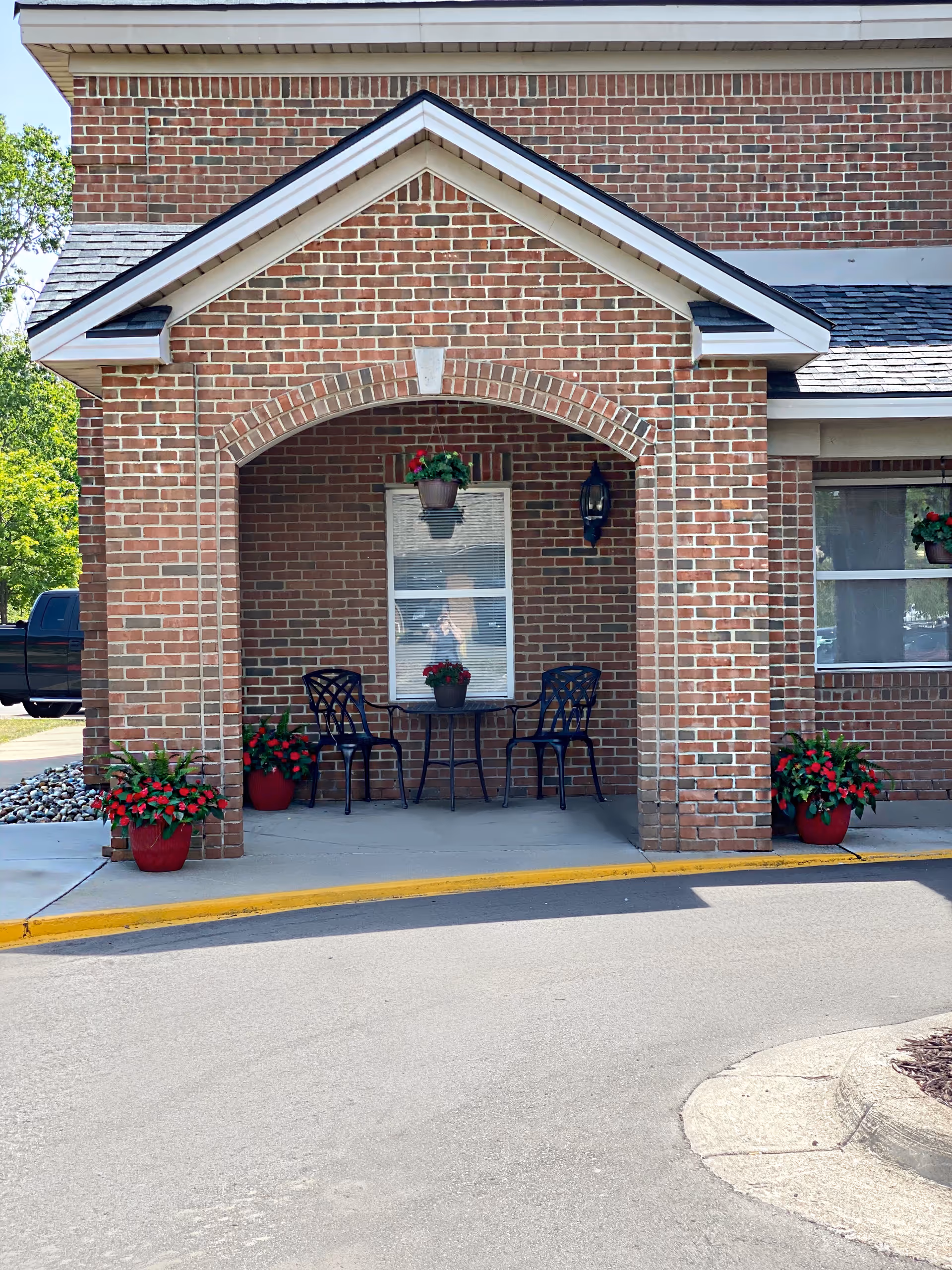 Brick exterior entrance of a building with an arched covered porch area containing a small round table and two black metal chairs. There are red flowering plants in pots placed around the porch and hanging from the ceiling. A window with blinds and a wall-mounted lantern light are visible on the brick wall behind the table.