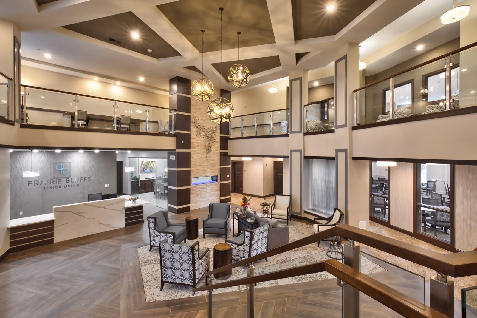 Spacious and modern senior living facility lobby with a high ceiling, decorative hanging lights, a stone accent wall with a built-in fireplace, and a reception desk with the Prairie Bluffs Senior Living logo on the wall. The seating area includes multiple chairs and small tables arranged on a patterned rug. There is a glass railing on the upper level overlooking the lobby.