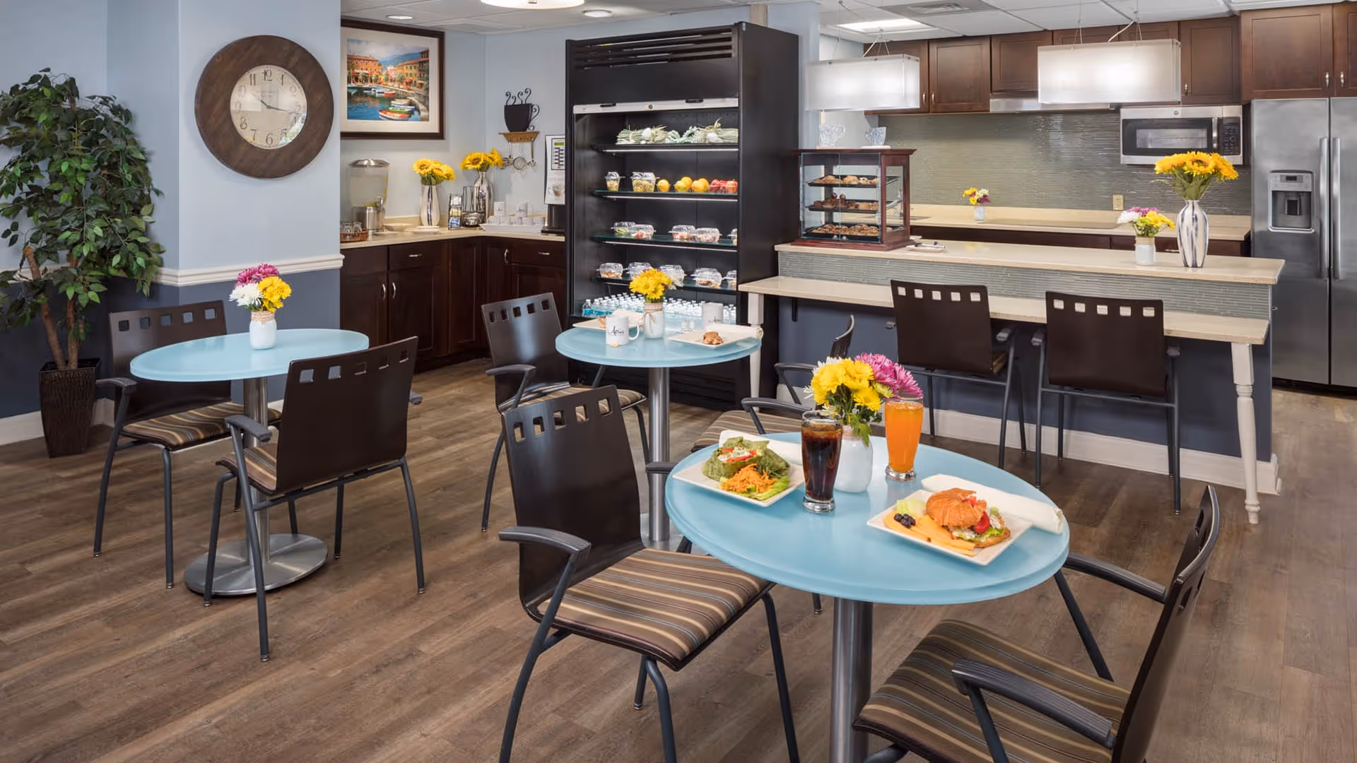 A cozy dining area in a senior living facility with light blue round tables and dark chairs with striped cushions. The tables have small vases with colorful flowers and plates of food with drinks. In the background, there is a kitchen area with a counter, bar stools, a refrigerator, microwave, and a display shelf with various food items. The room has wooden flooring, light blue walls, a large clock, and a potted plant in the corner.