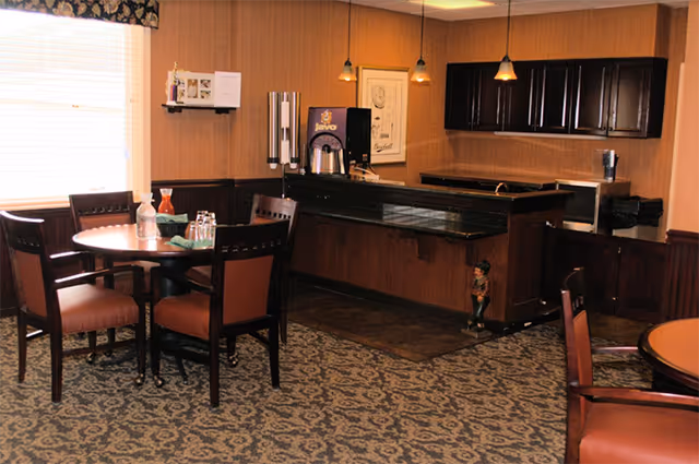 Interior view of a dining area in a senior living facility featuring round wooden tables with chairs, a coffee and beverage station with a coffee maker and cups, pendant lights hanging from the ceiling, and a patterned carpeted floor.