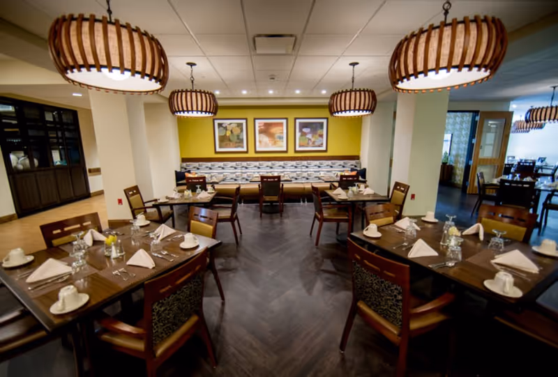 Well-lit dining room with neatly set tables, pendant lights, and banquette seating against a green accent wall.