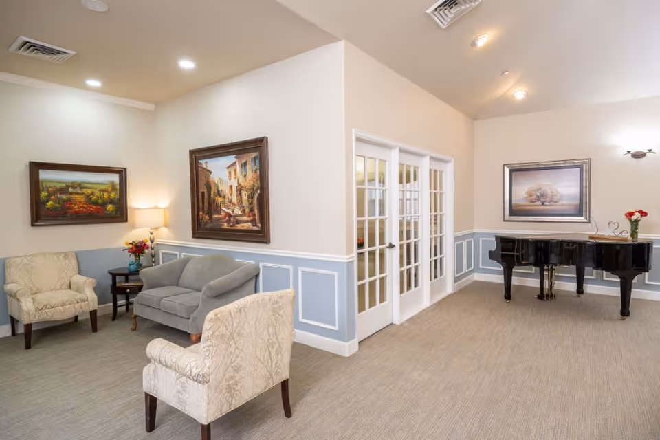 Bright seating area in a senior living common room with upholstered chairs, a small sofa, framed paintings, French doors, and a grand piano.