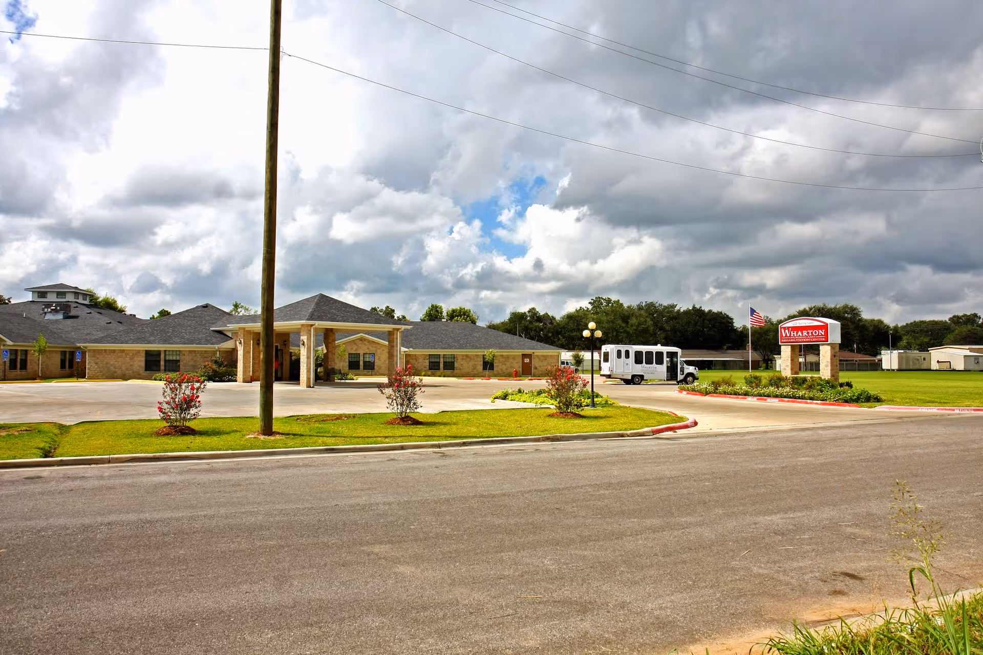 Front view of Wharton Nursing and Rehabilitation Center with a covered entrance, driveway and parked shuttle van under a cloudy sky.
