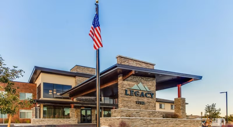 Exterior view of Legacy House of Southern Hills building with a stone facade, large windows, and a covered entrance. An American flag is prominently displayed on a flagpole in front of the building under a clear blue sky.