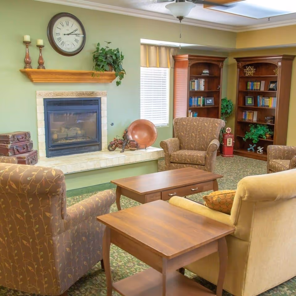 Cozy common living room with upholstered armchairs and wooden coffee tables arranged around a fireplace and bookshelves.
