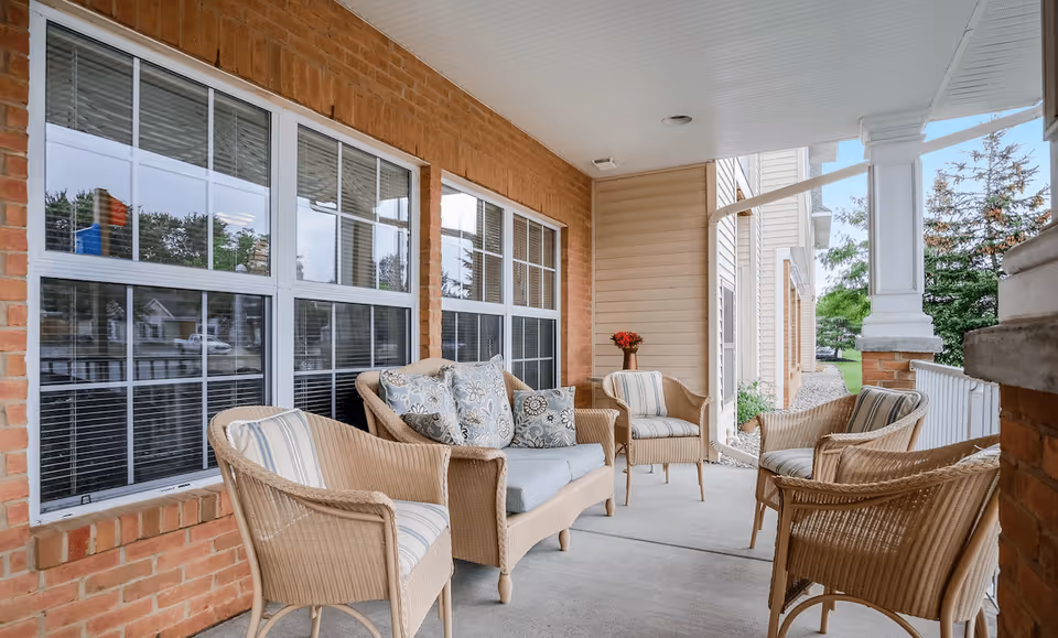 Covered outdoor patio area with wicker furniture including a loveseat with cushions and four armchairs arranged around a small table with a vase of flowers. The patio has brick walls and large windows, with a view of greenery and trees outside.