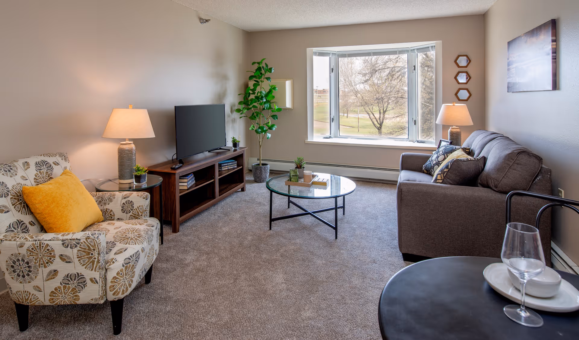 Cozy living room with an armchair, sofa, TV on a wooden stand, round glass coffee table, lamps and a large bay window overlooking trees.