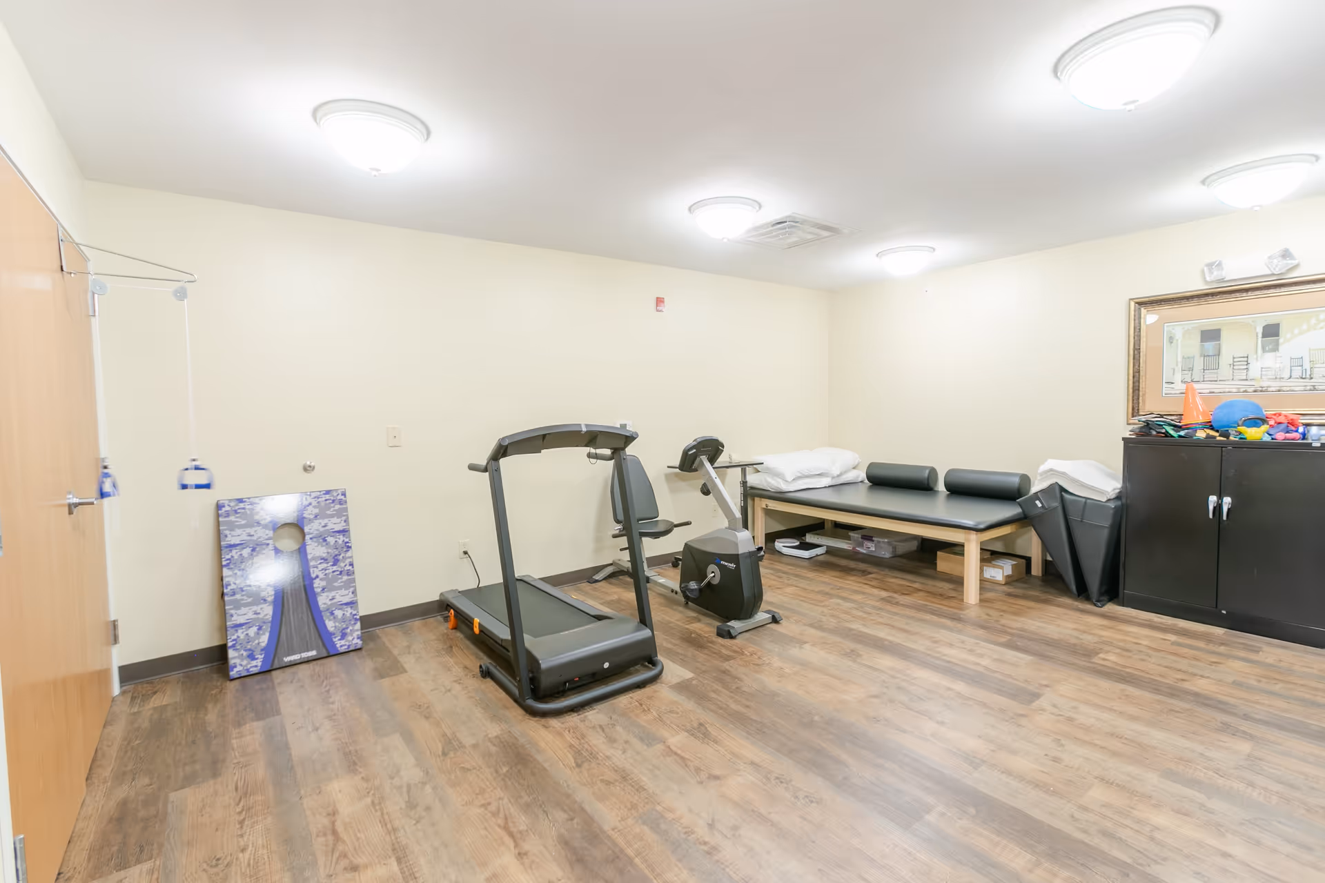 A small exercise room with a treadmill, stationary bike, therapy table with pillows, a cabinet with exercise equipment, and a cornhole board leaning against the wall. The room has wood flooring, cream-colored walls, and ceiling lights.