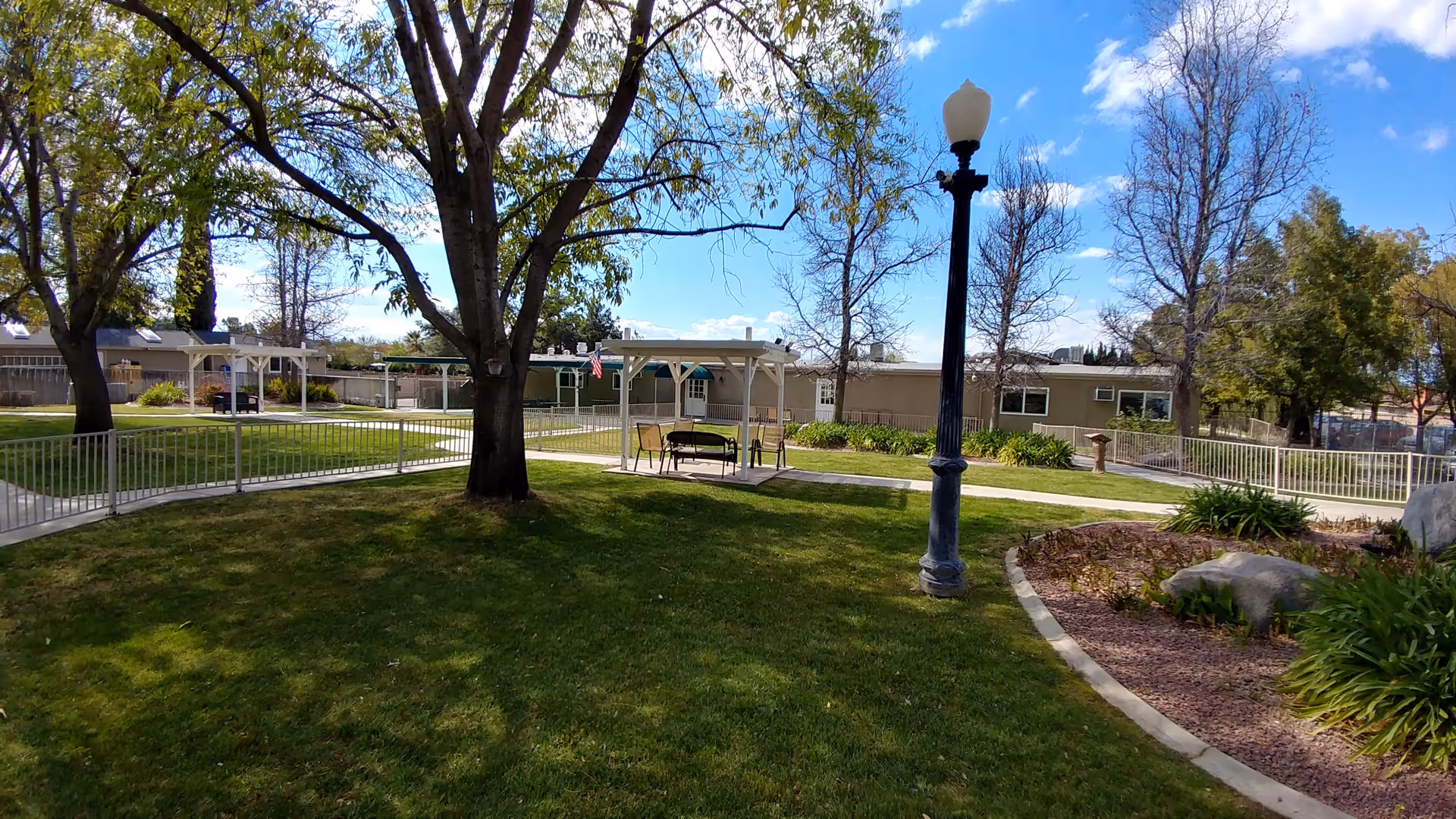Outdoor garden area at Rose Garden Residential Care with green grass, trees, a lamppost, paved walkways, benches under small pergolas, and a clear blue sky with some clouds.