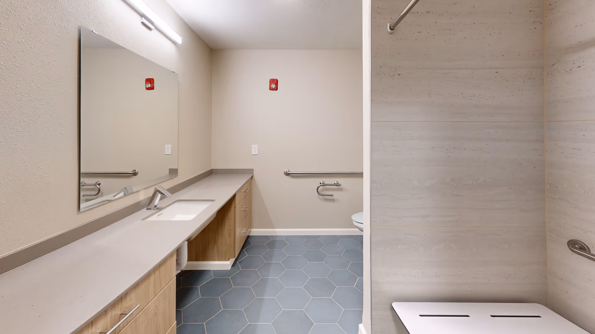 Accessible bathroom with a long countertop and sink, hexagonal tile floor, grab bars, toilet and a shower bench.