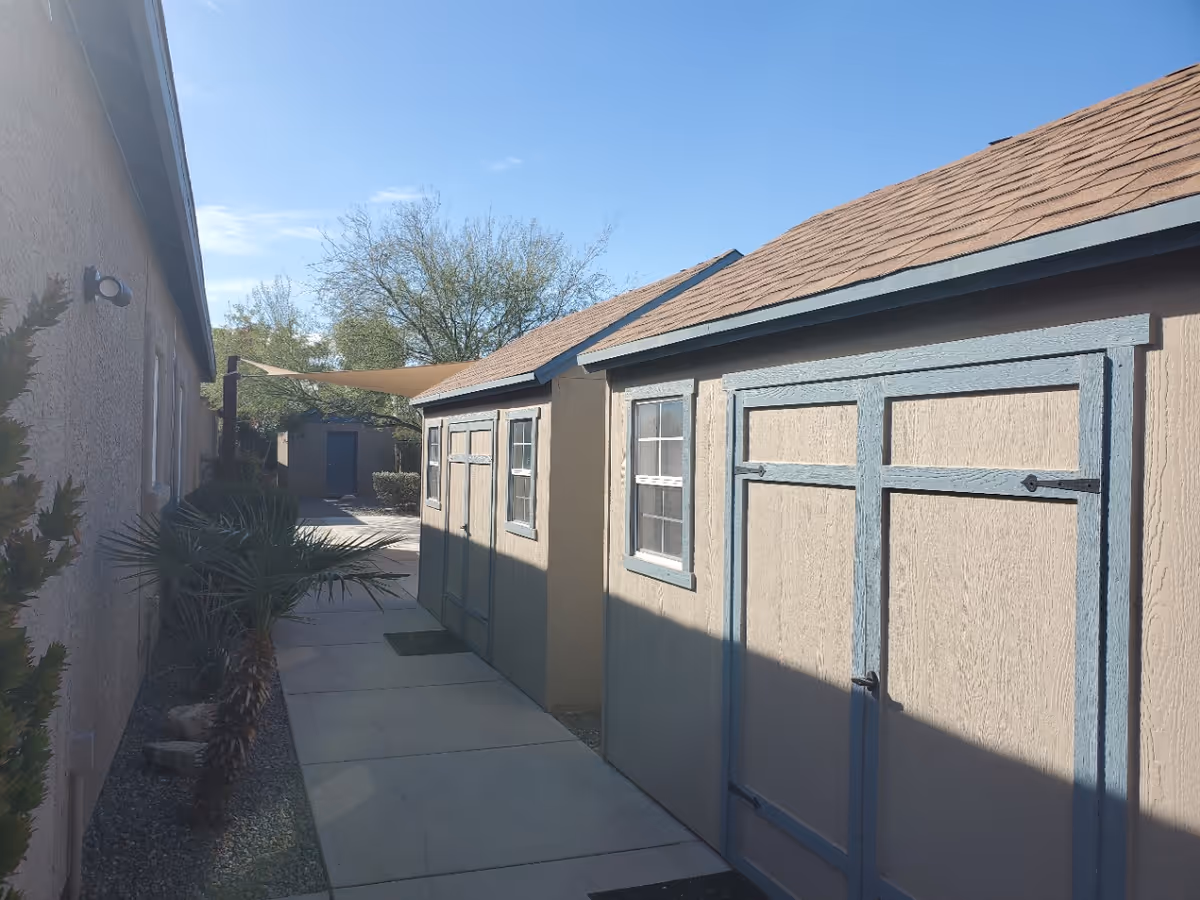Outdoor walkway between a building and two small storage sheds with beige walls and blue trim under a clear blue sky with some trees in the background.
