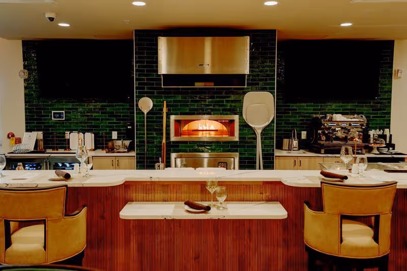 Bar-style dining counter with place settings and chairs facing a tiled wall with a lit pizza oven and coffee machine behind.