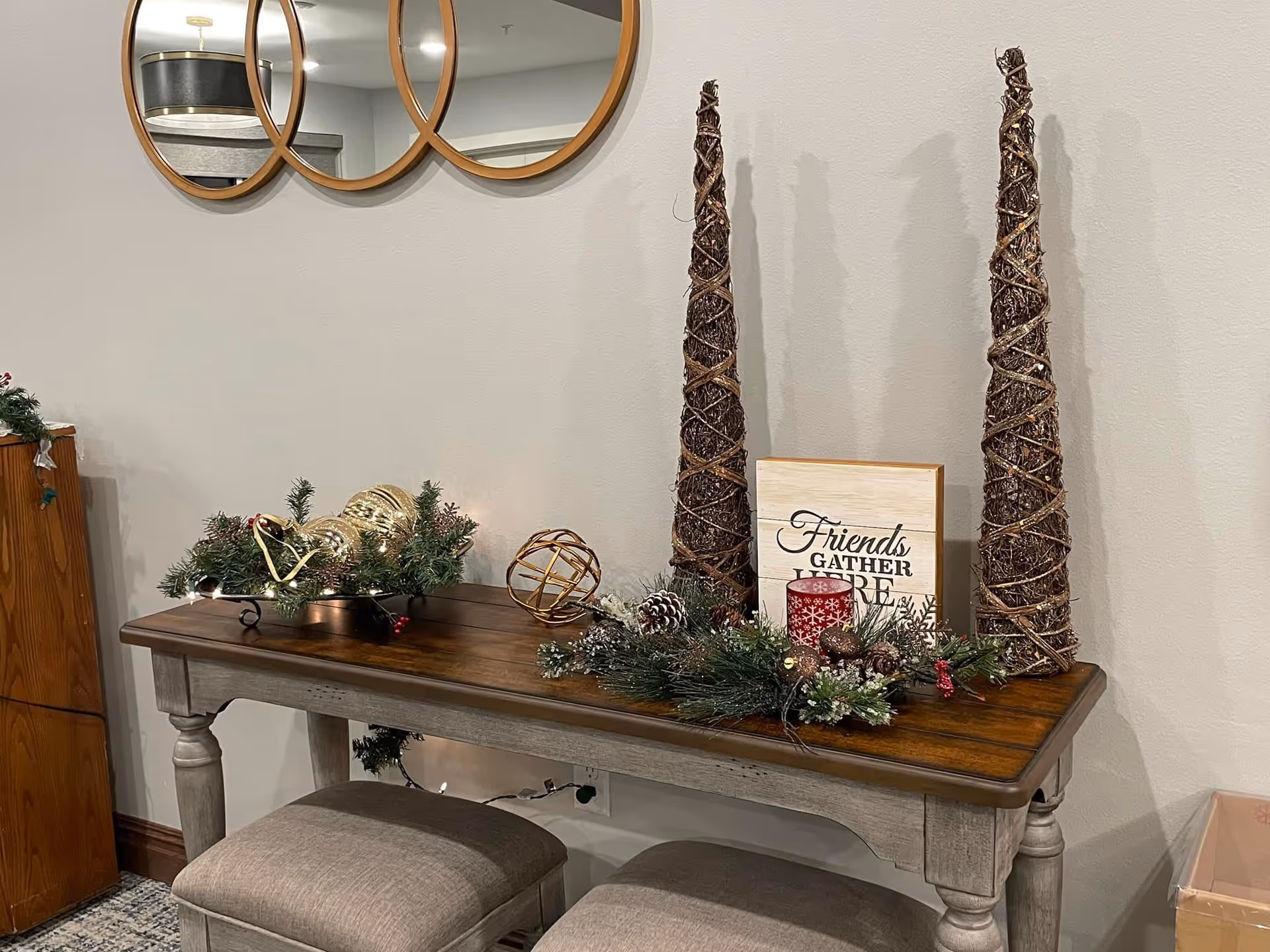 A wooden console table with a dark wood top and light gray legs, decorated with holiday greenery, pinecones, gold ornaments, a red candle, and two tall cone-shaped twig decorations. Above the table are three round mirrors with wooden frames mounted on a light gray wall. Two cushioned stools are placed under the table.