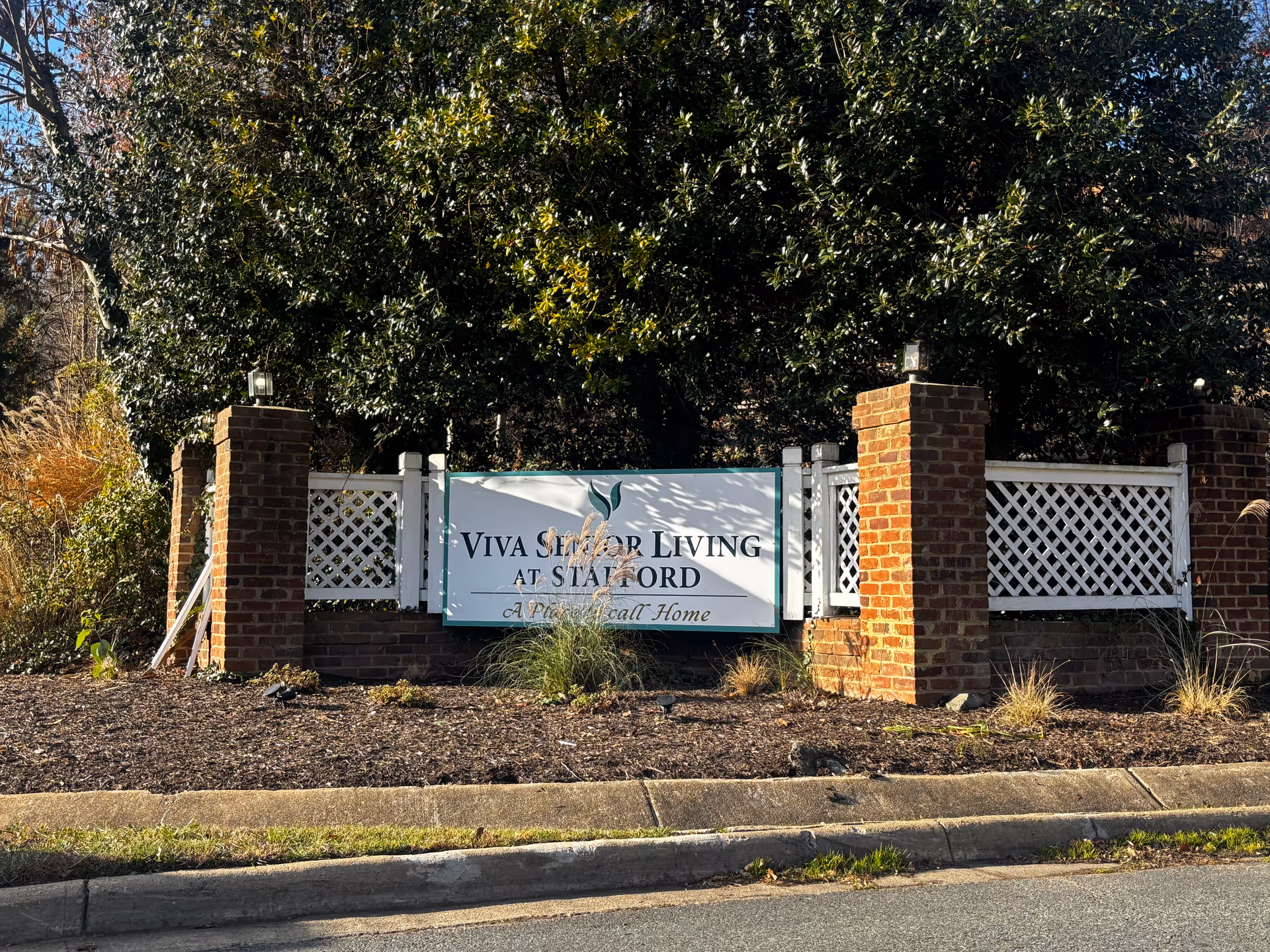 Entrance sign reading 'Viva Senior Living at Stafford' mounted between brick pillars and white lattice fencing in front of shrubs.