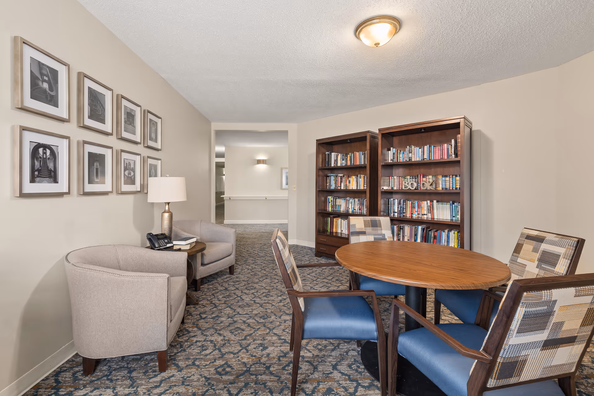 A cozy senior living common area with a round wooden table surrounded by four chairs with patterned upholstery. Two wooden bookshelves filled with books are against the wall. To the left, there are two beige armchairs with a small round table between them holding a lamp and a telephone. Several framed black and white photos hang on the wall above the armchairs. The carpet has a blue and beige pattern, and the walls are painted light beige.